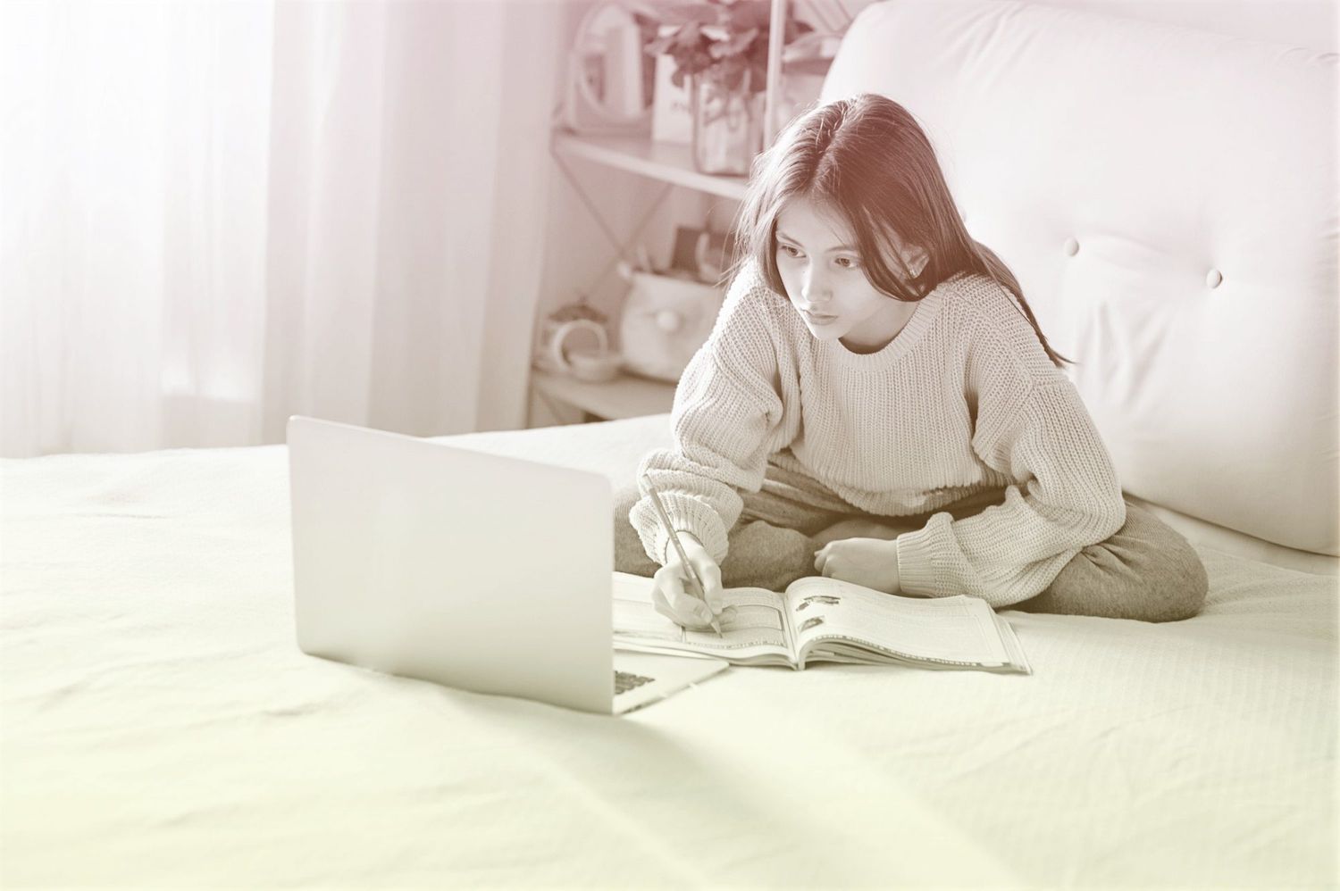 young girl doing schoolwork on bed with laptop and book