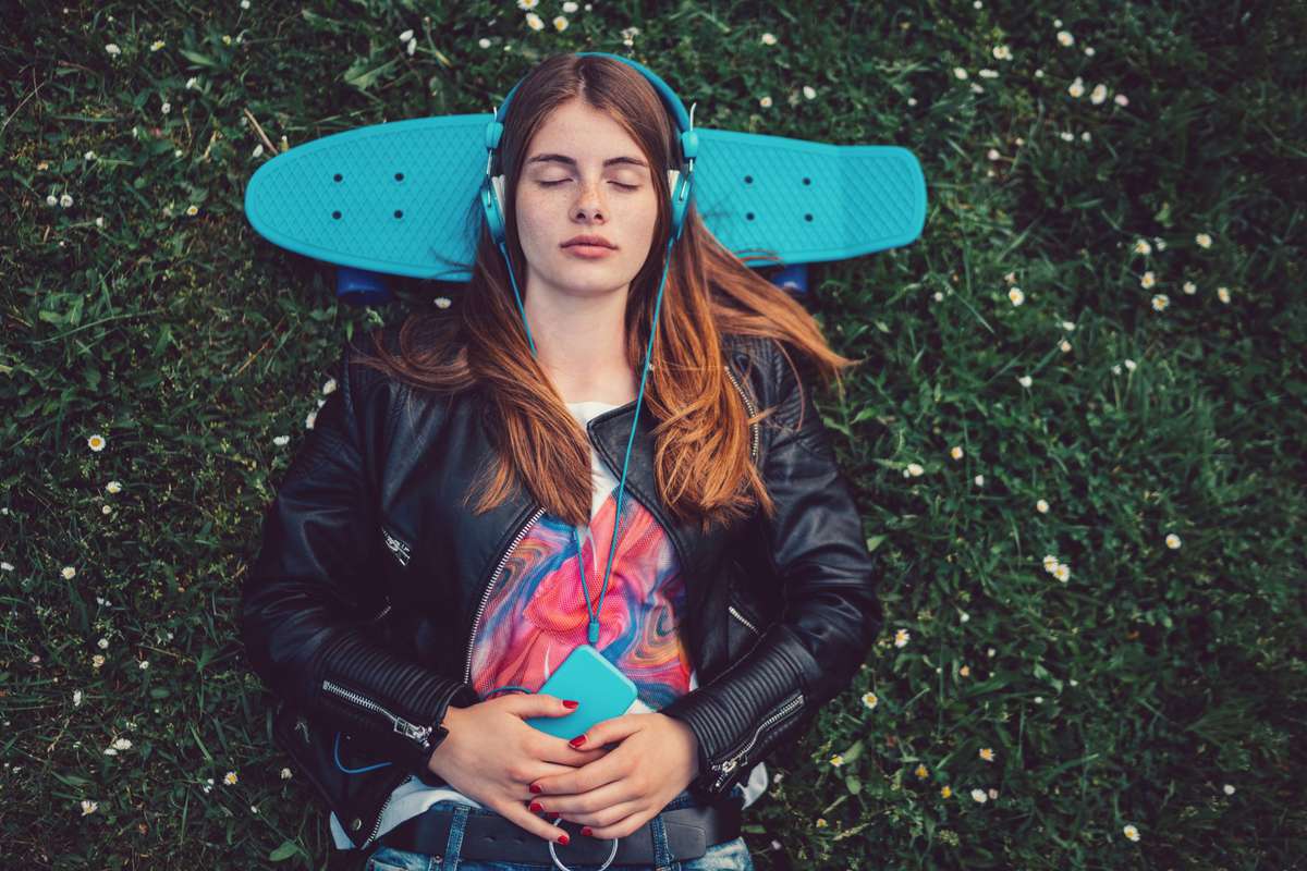 Relaxed teenage girl in the grass listening to music - Young woman lying down in the grass with skateboard behind head