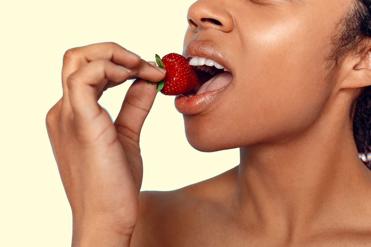 best-wrost-foods-teeth , Young african woman with bare shoulders standing isolated on blue background holding strawberry biting sensual mouth close-up