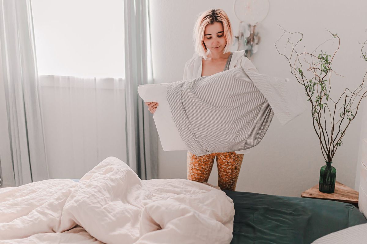 How Often Should You Wash Your Pillowcases? , Young pretty woman is making a bed. Fresh bedding linen. Put on pillow cover. Light and bright room. Beige, white, grey. Doing home improvement. Cleaning and organizing. Cozy house. Apartment living.