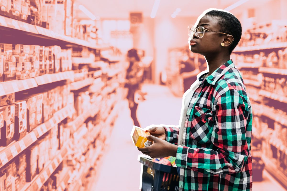 一位年轻女子提着一篮子商品在超市过道里寻找食物。