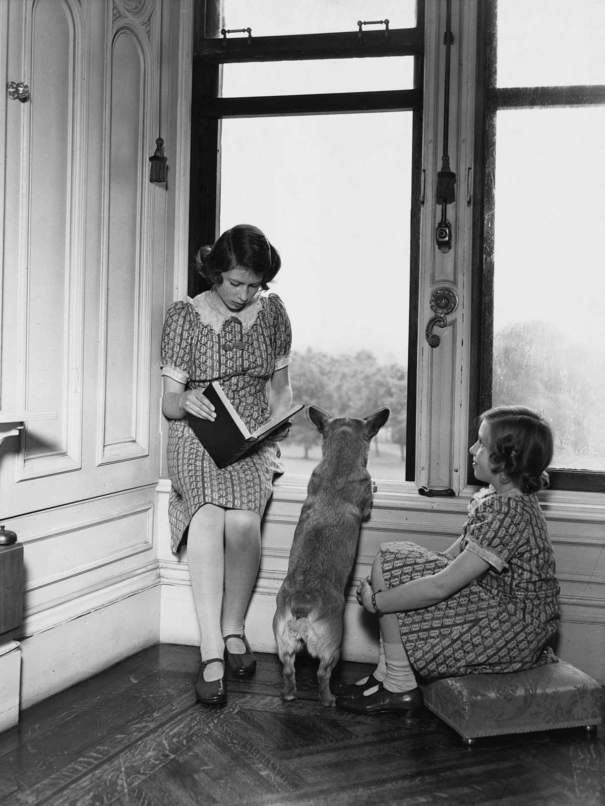 Queen Elizabeth reads to her sister, while her corgi looks out the window at Windsor Castle