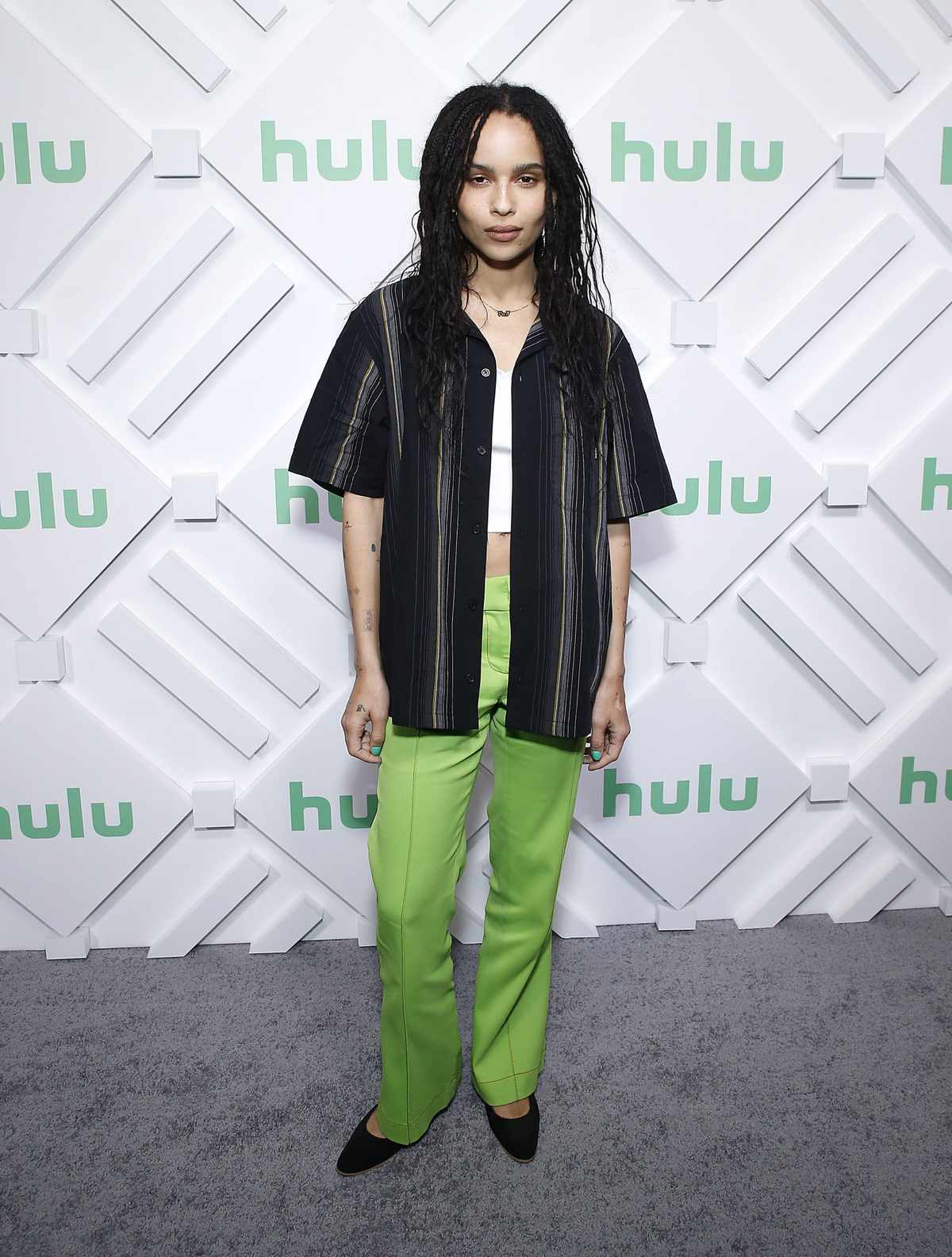 Zoe Kravitz wearing green pants, a black striped short sleeve button down, and a white tank top with black shoes