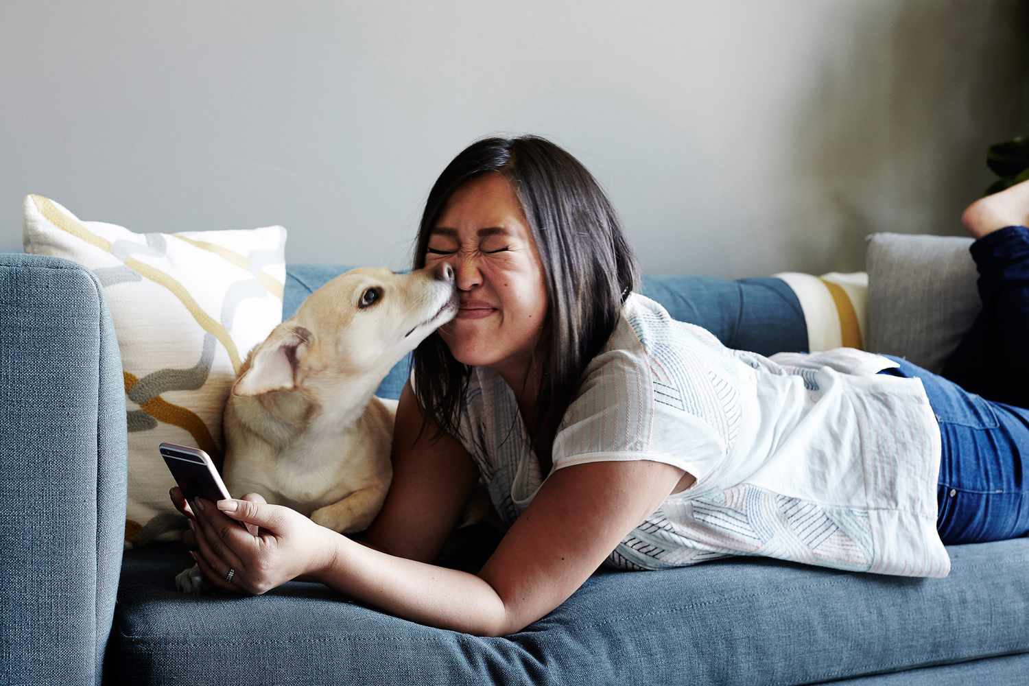Woman lying on sofa using smartphone, pet dog licking her face