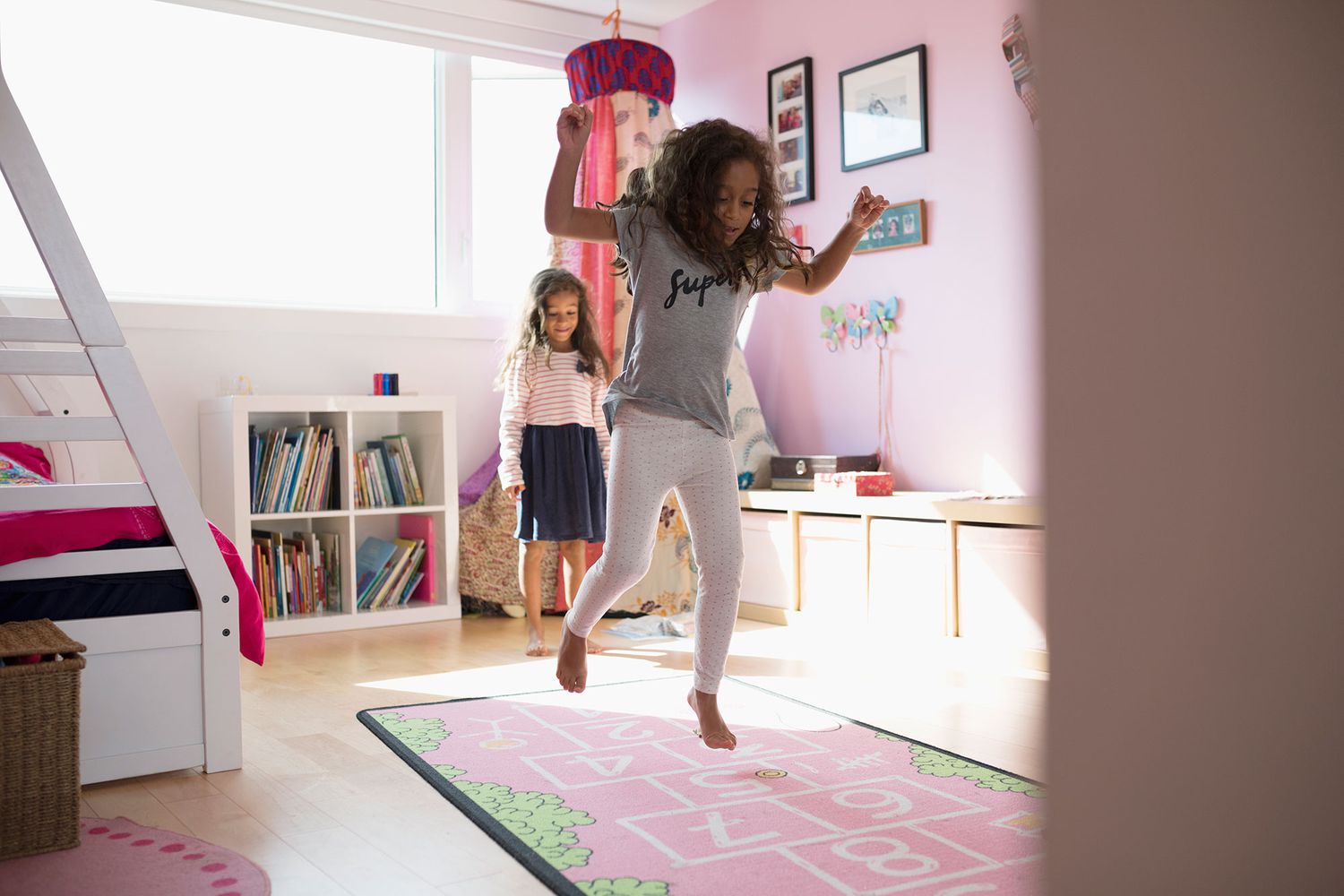 Girl sisters playing hopscotch on pink rug in bedroom