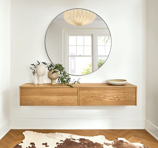 floating wooden cabinet with two drawers under a circle mirror on a white wall, wooden floor with a cow-print rug