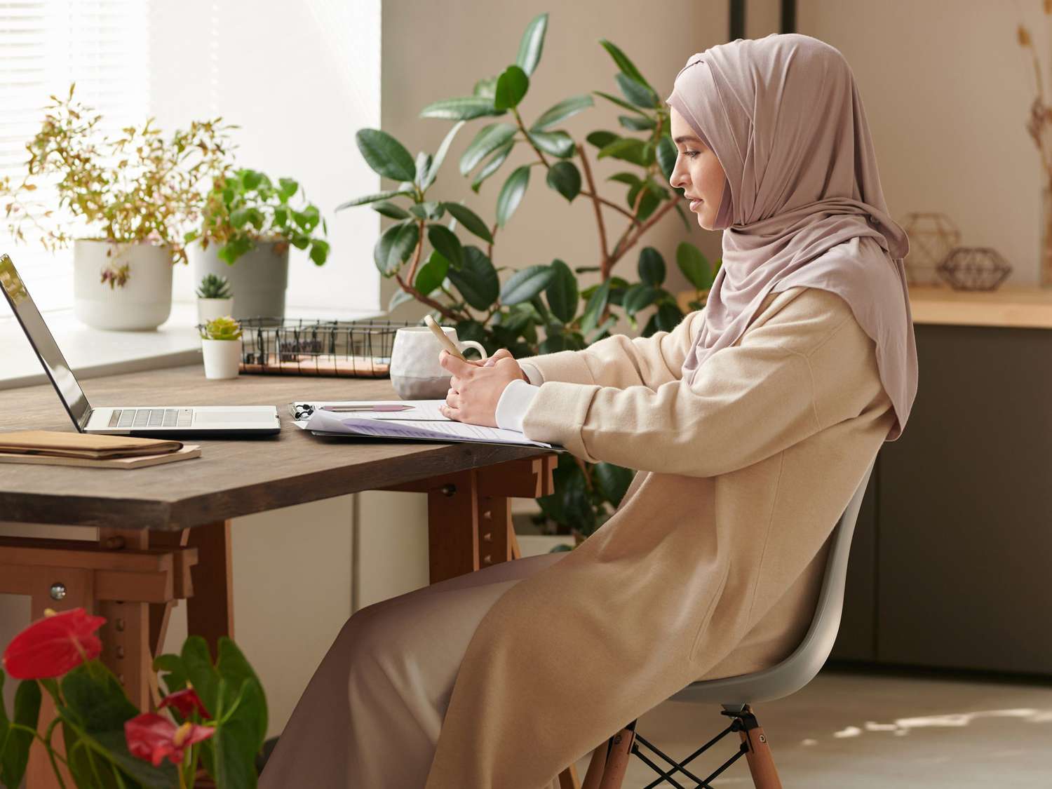 Young Muslim woman wearing headscarf sitting alone in cozy office room surfing Internet on smartphone during break, copy space