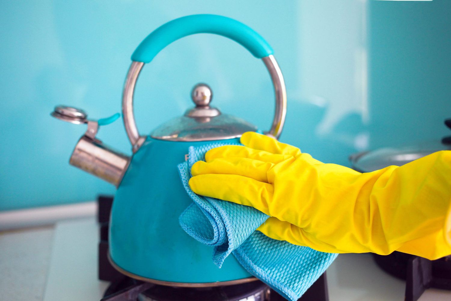 A female hand in a rubber yellow glove wipes a blue teapot with a rag.