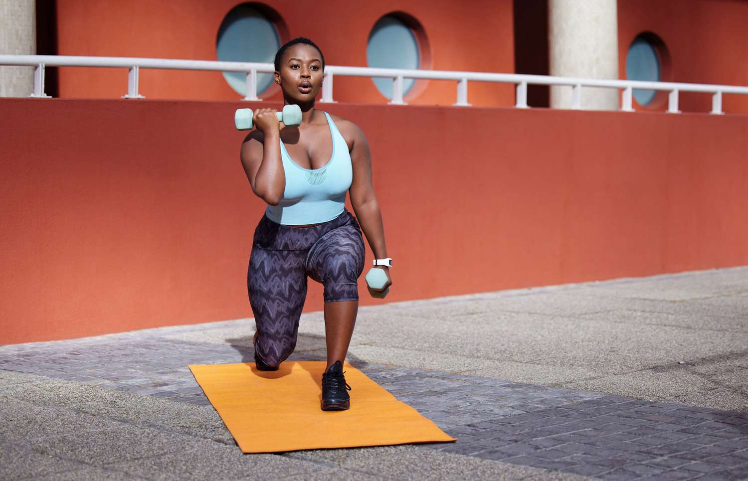 Shot of a young woman on a gym mat using dummbells against an urban background