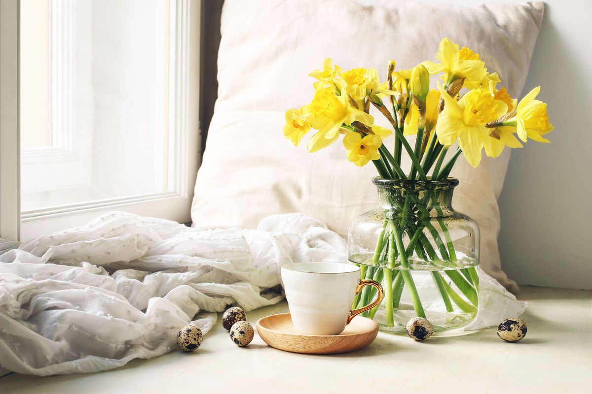 Cup of coffee, wooden plate, quail eggs and vase of flowers on windowsill. Floral composition with yellow daffodils, narcissus.