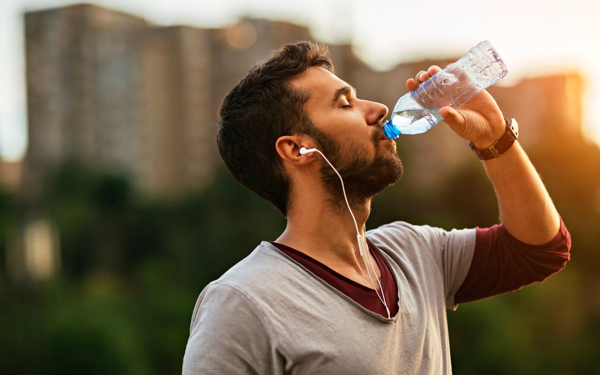 Close up of a young man drinking water from a plastic bottle . He is listening to music from small white earphones and his eyes are closed. The sun is shining strong in the background.