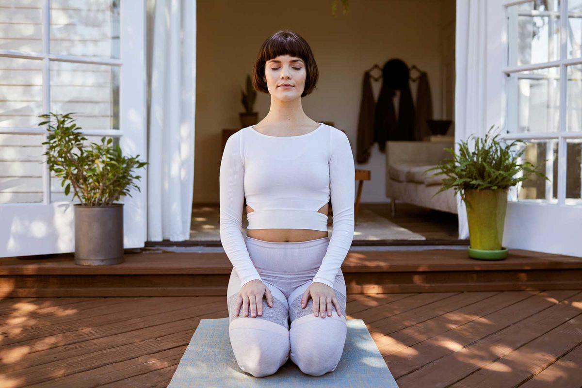Young woman in sportswear kneeling on an exercise mat on her patio and meditating with her eyes closed during meditation