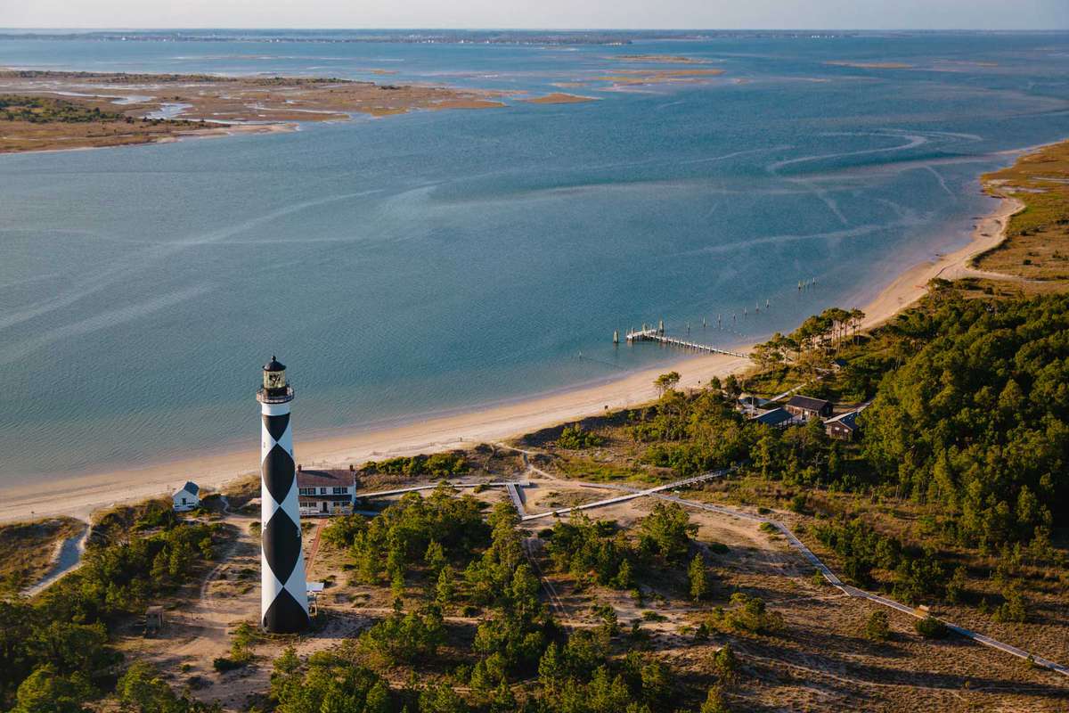 Aerial view of Cape Lookout National Seashore, Cape Lookout Lighthouse and Keepers' Quarters Museum, National Park Service offices, gift shop and dock with Barden's Inlet, Shackleford Banks and Harker’s Island in the distance and green foliage along the shoreline.