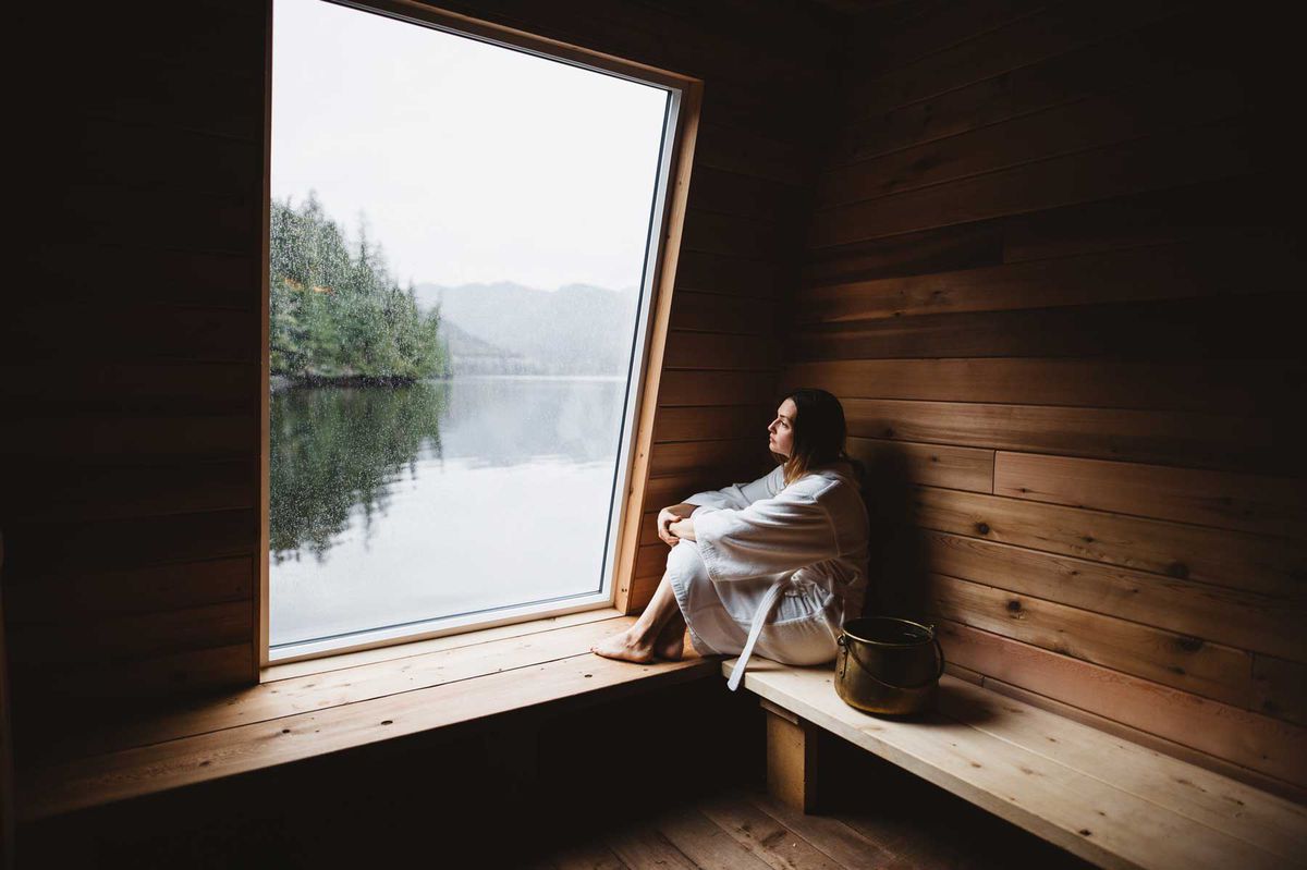 Woman sitting in sauna staring out of the window at water and trees