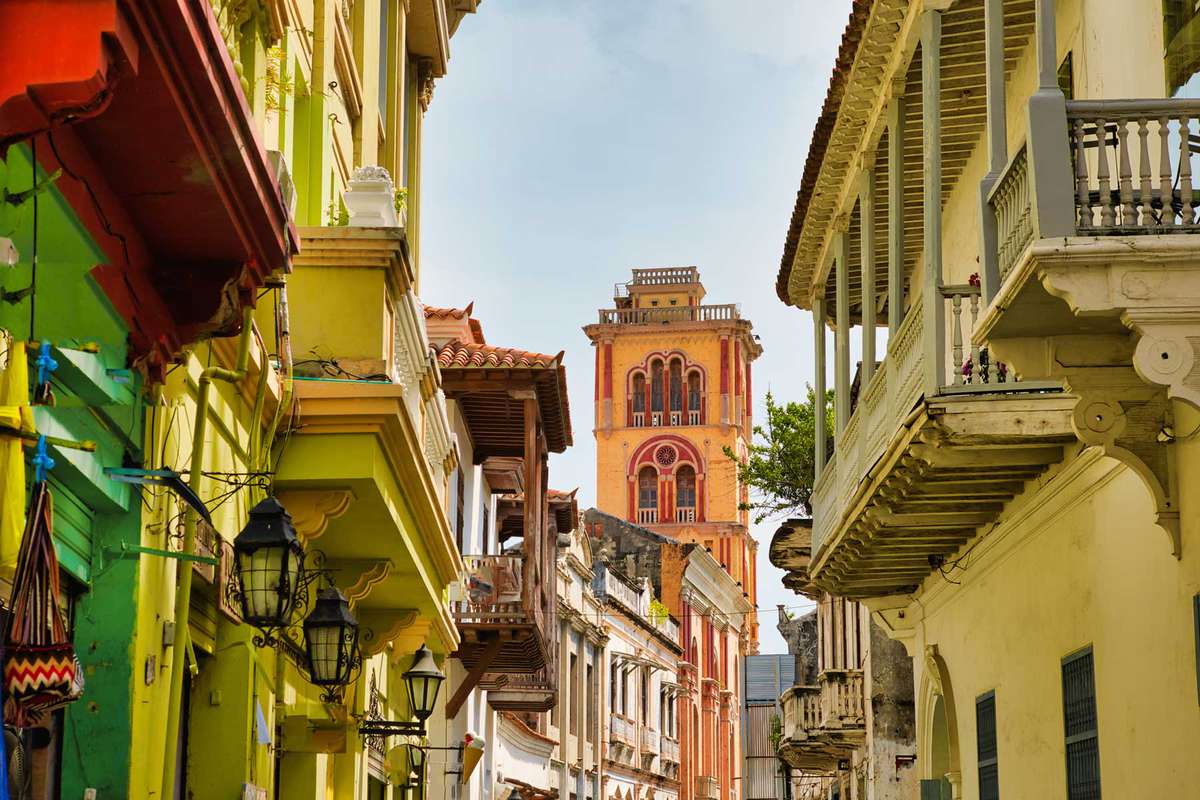 Spanish colonial houses in vivid assorted colors, lined up. Beautiful architectural details are noted throughout the buildings, as well as charming verandas. In the distance, a sturdy tower stands out against the soft blue sky.