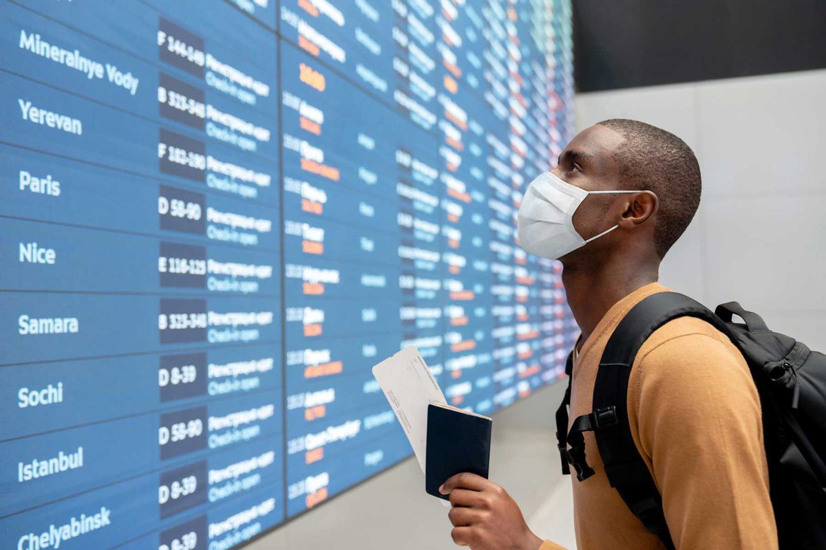 Portrait of a black male traveler wearing a facemask at the airport and looking at the flight schedule