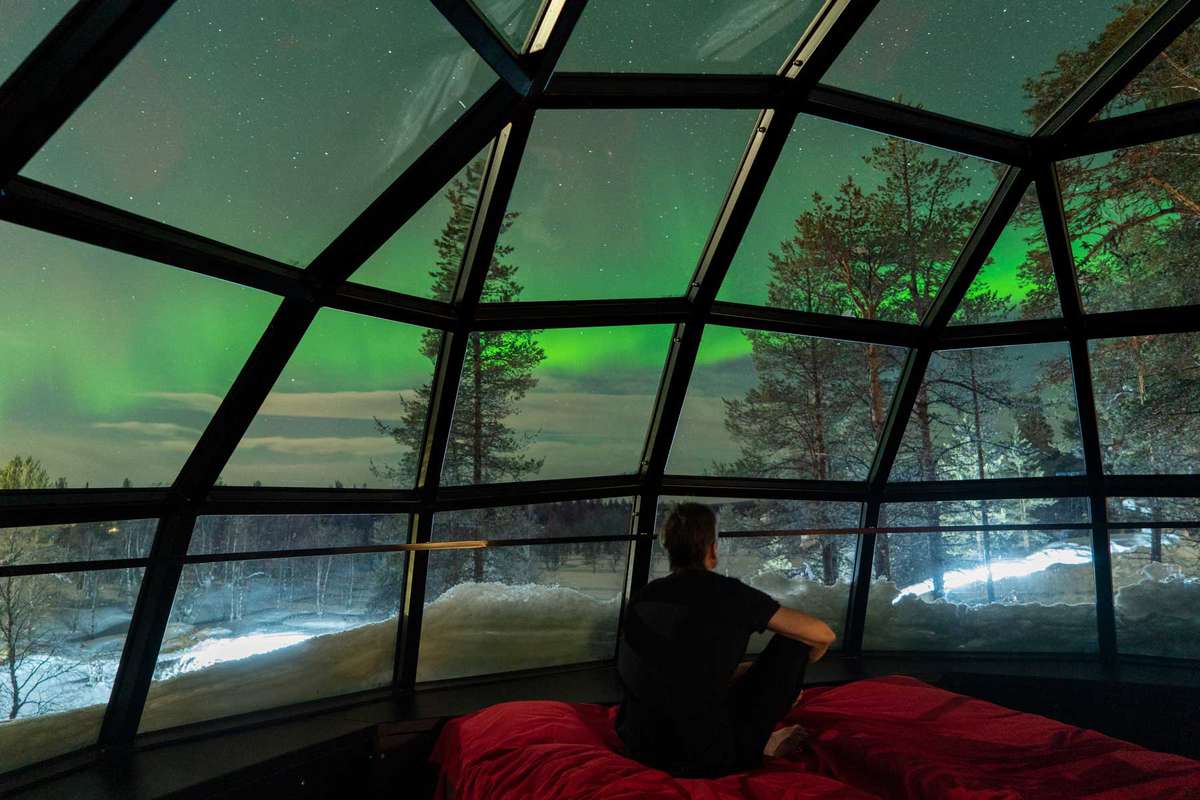 A man sits on a red bed in a glass guest tent in Finland, observing the green sky (Northern Lights) and stars