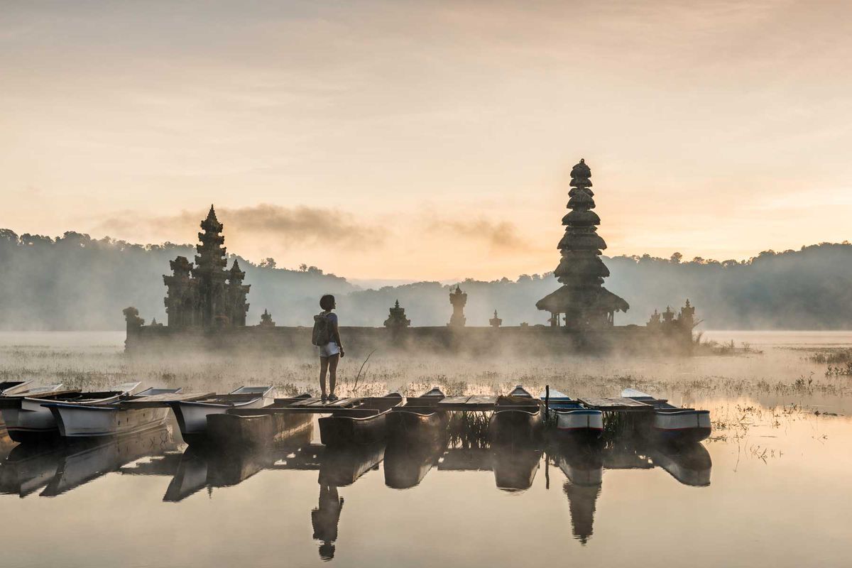 Young Asian woman standing on boat admiring Tamblingan temple, submerged in water, with early morning mist shrouding the Hindu temple.