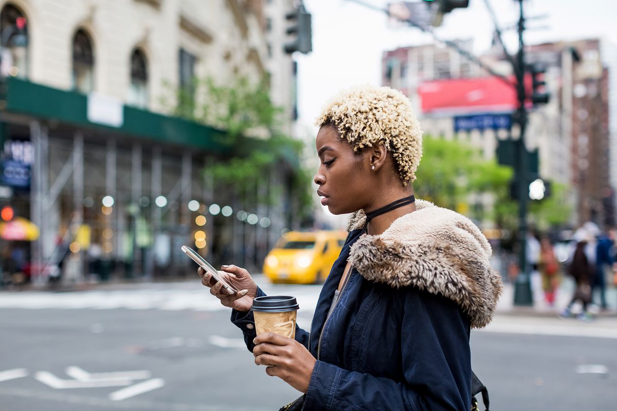 A fashionable young woman with coffee to go looking at cell phone on the street