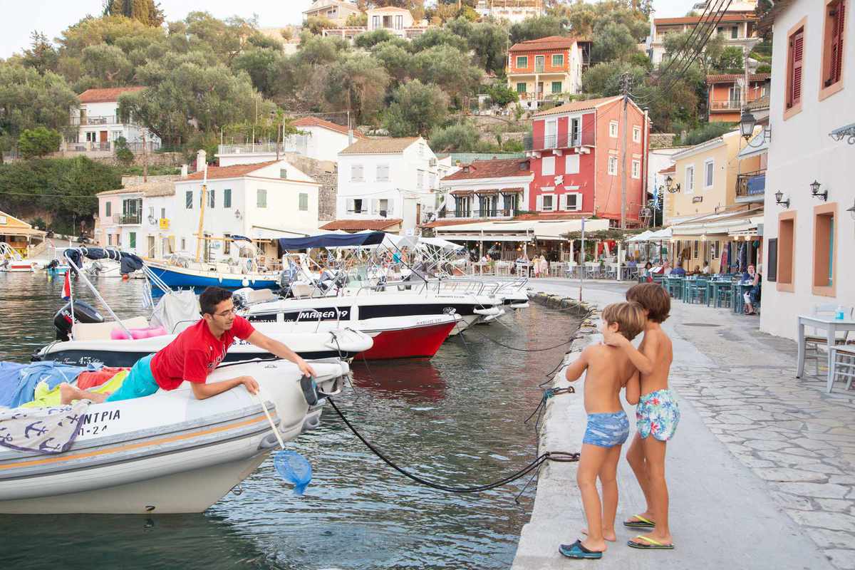 Two kids standing by a Greek harbour, with a man anchoring his boat