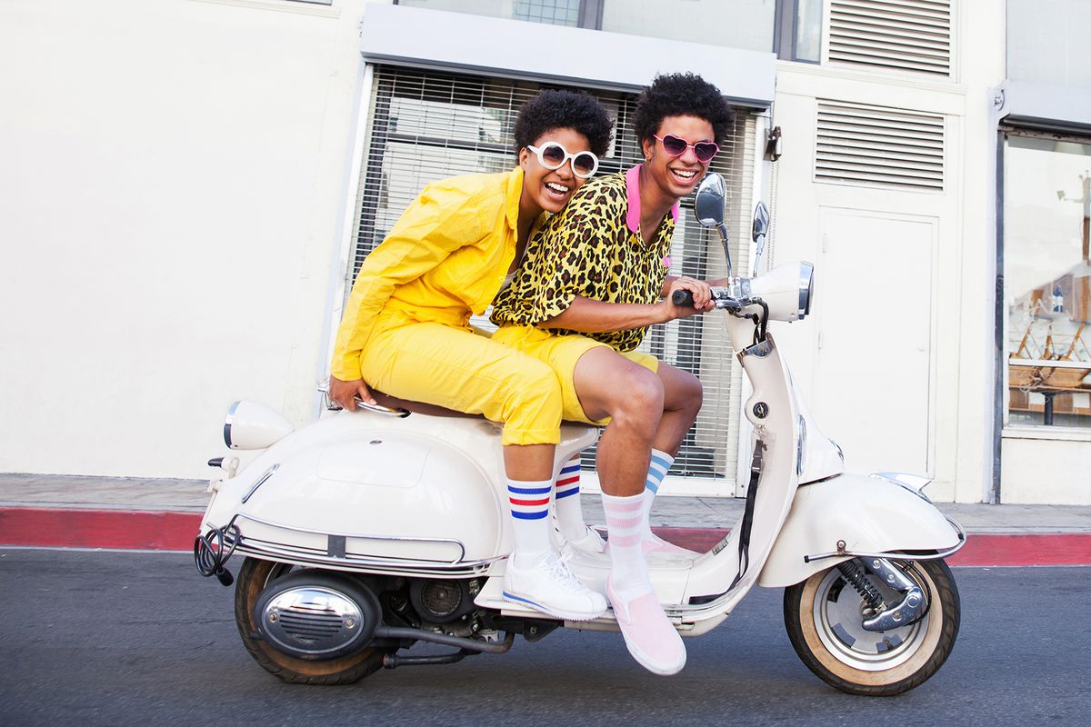 A hip young couple wearing yellow outfits and large sunglasses riding a scooter on a city street looking at camera smiling
