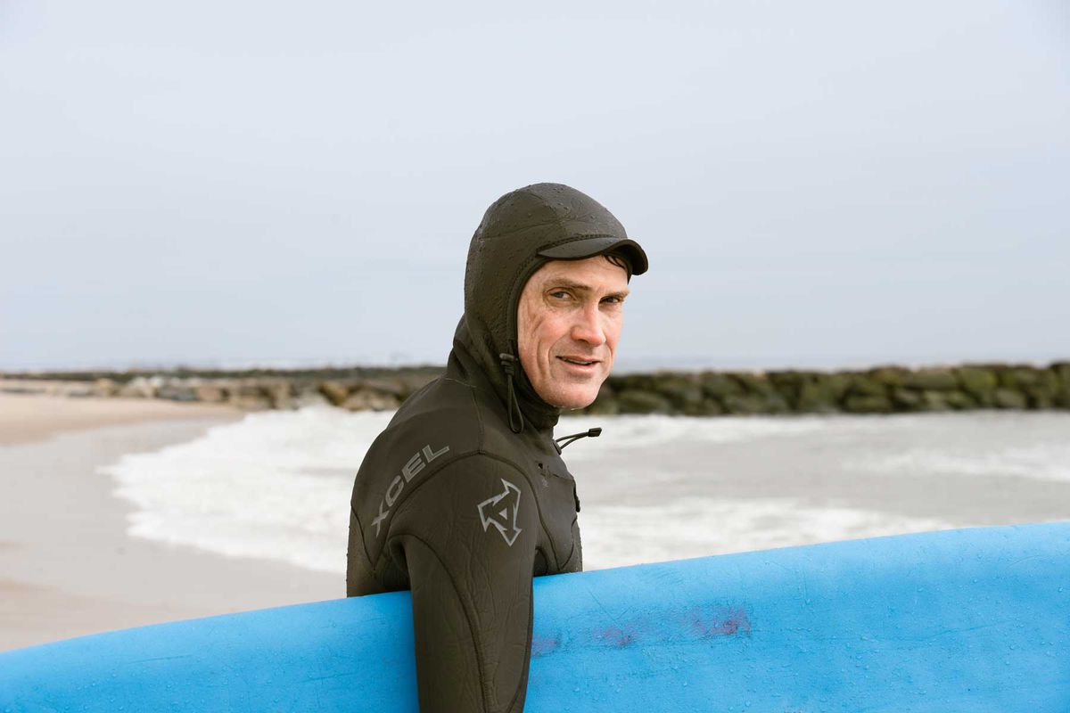 Writer Tom Vanderbilt wearing a full body wetsuit and holding a blue surfboard