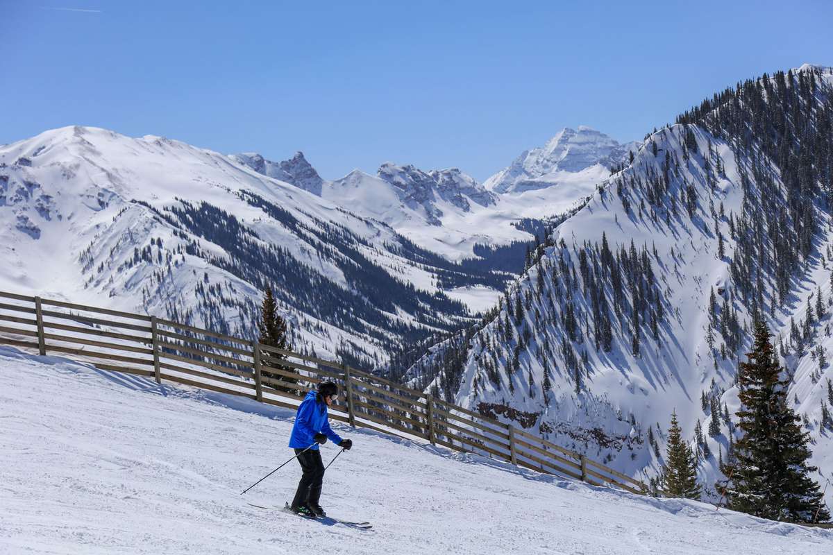 A man in a blue jacket downhill skiing with the Maroon Bells of the Rocky Mountains in the background at Aspen Snowmass ski resort