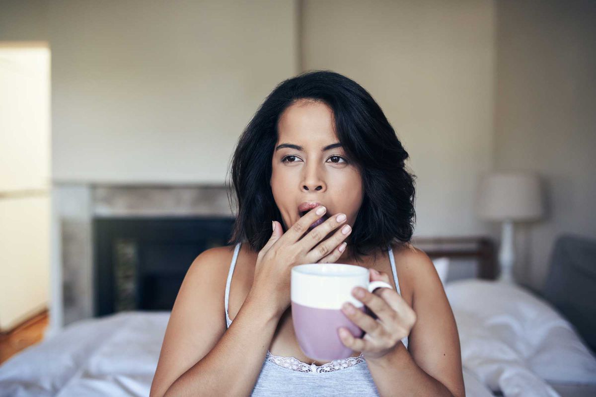 Shot of a young woman yawning while having coffee in the morning at home