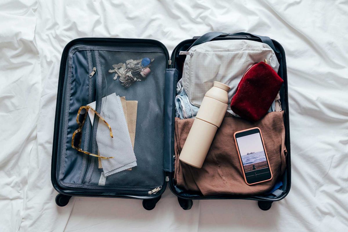 A packed suitcase from above on the bed on a holiday or a business trip containing a water bottle, an unlocked smartphone, a cosmetic bag, sunglasses, plane tickets, clothes and keys