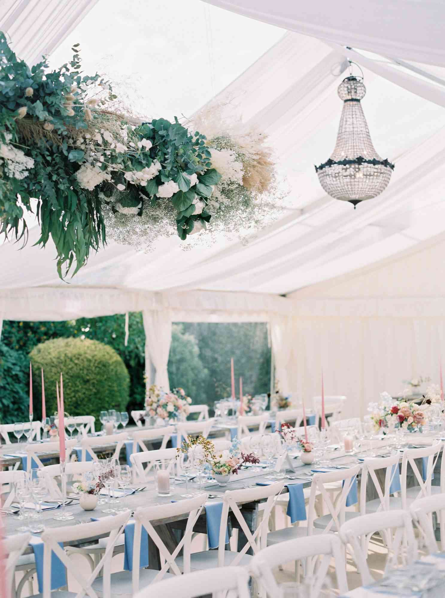 tented reception with long wooden tables, white chairs, chandeliers, and hanging floral arrangements