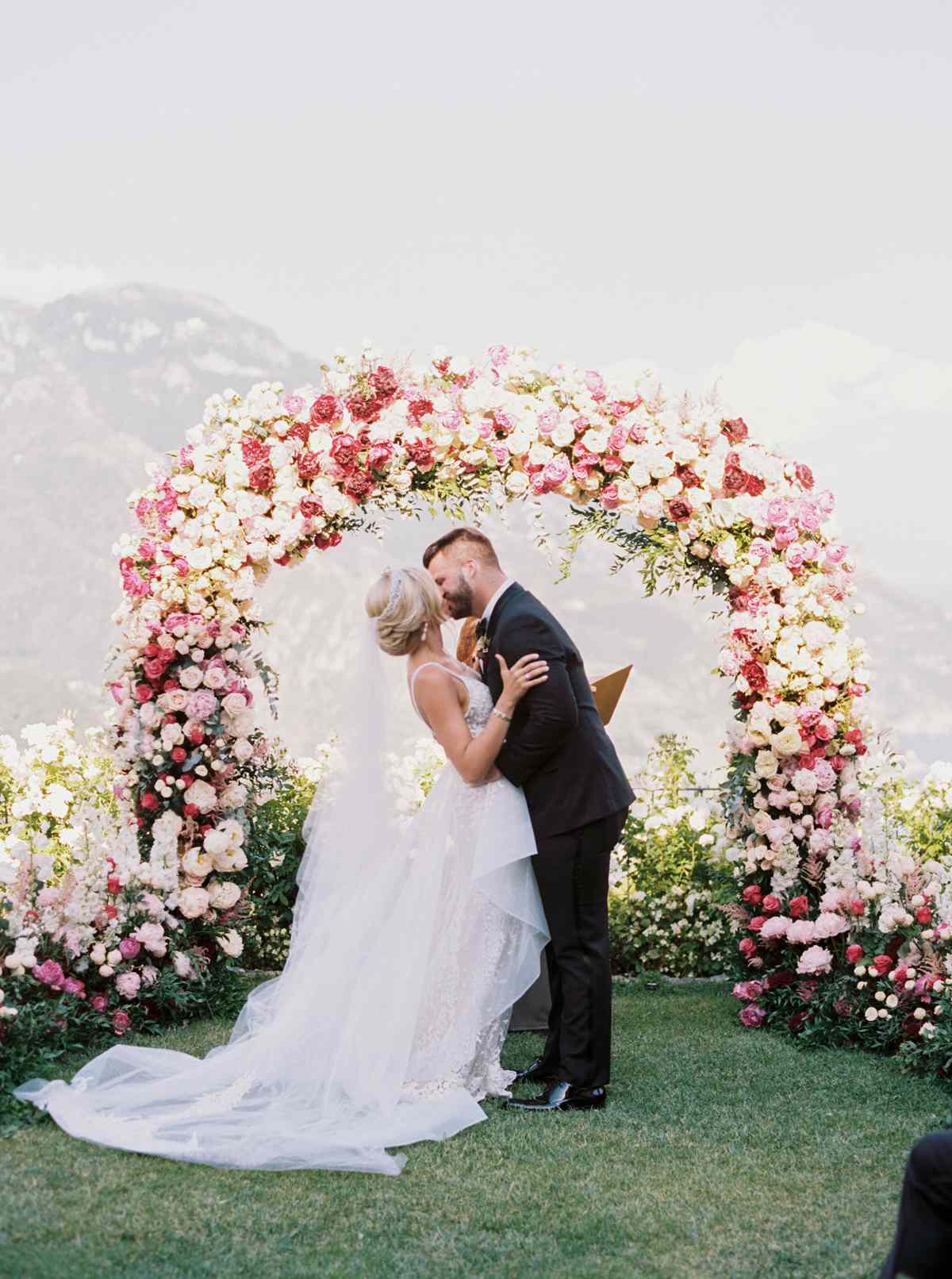allie and joe italy wedding couple kissing beneath floral arch