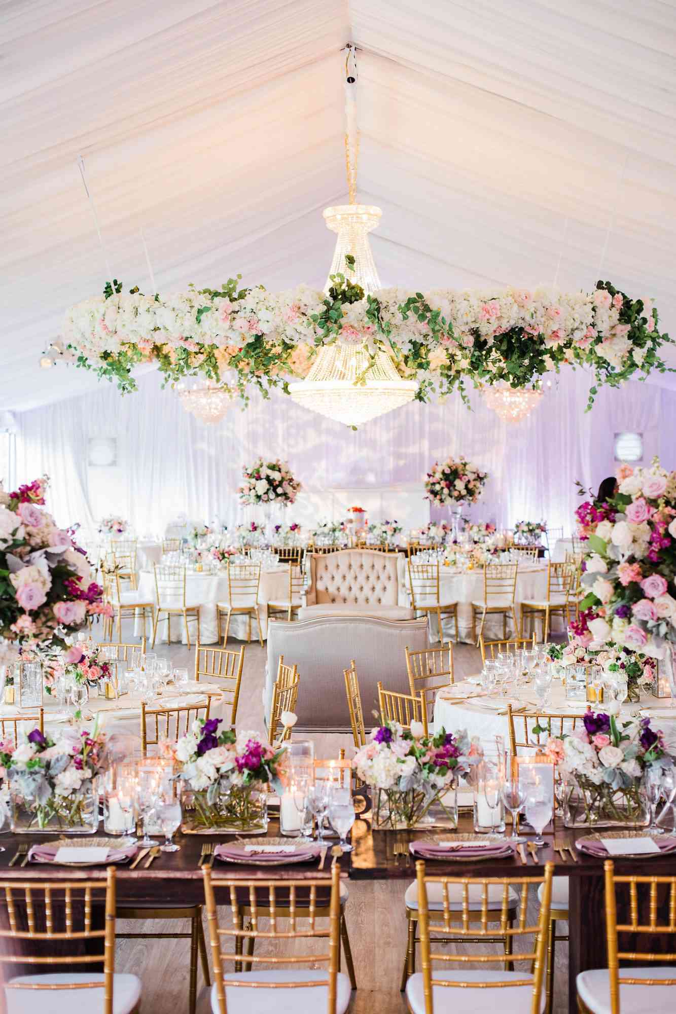 white tent wedding reception filled with hanging floral displays, centerpiece arrangements, and white and dinning furniture