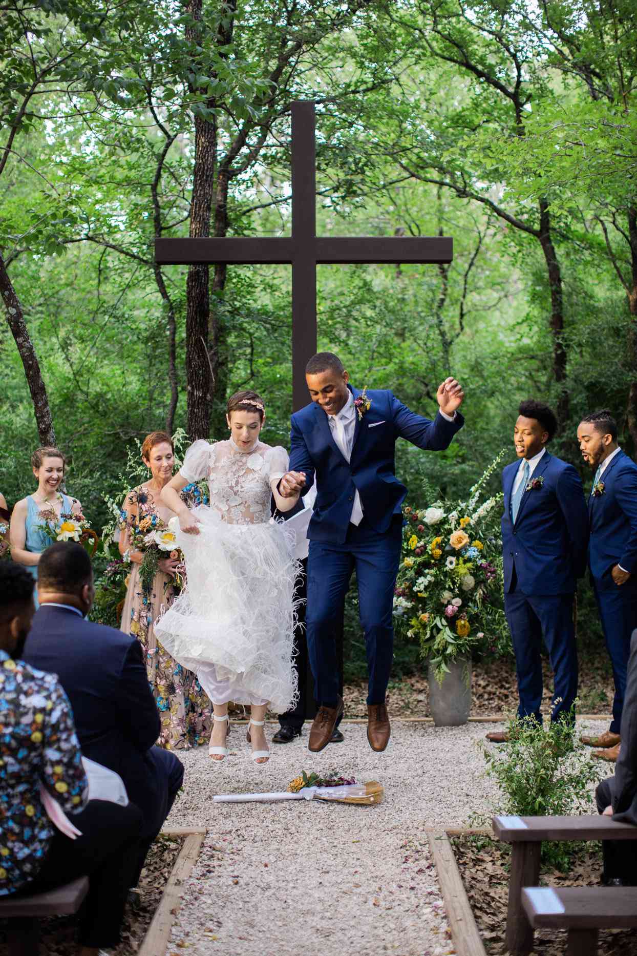Bride and groom jumping over a broom while wedding party and guests watch