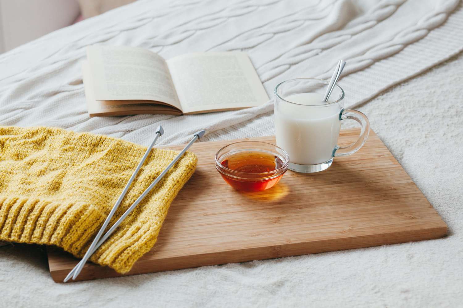 Knitting needles and yarn alongside hot milk in a glass cup and honey on a wooden board.