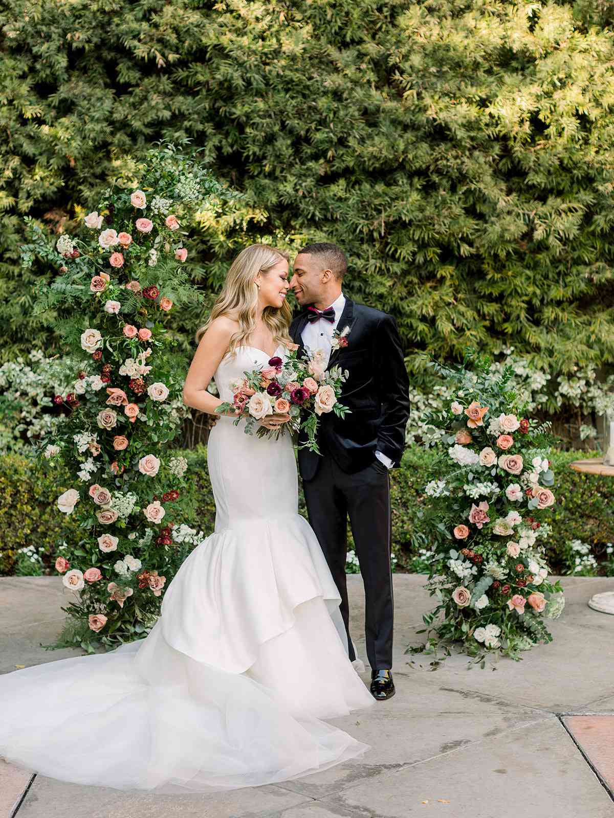 bride in white mermaid wedding dress and groom in black posing for portrait in front of floral arch