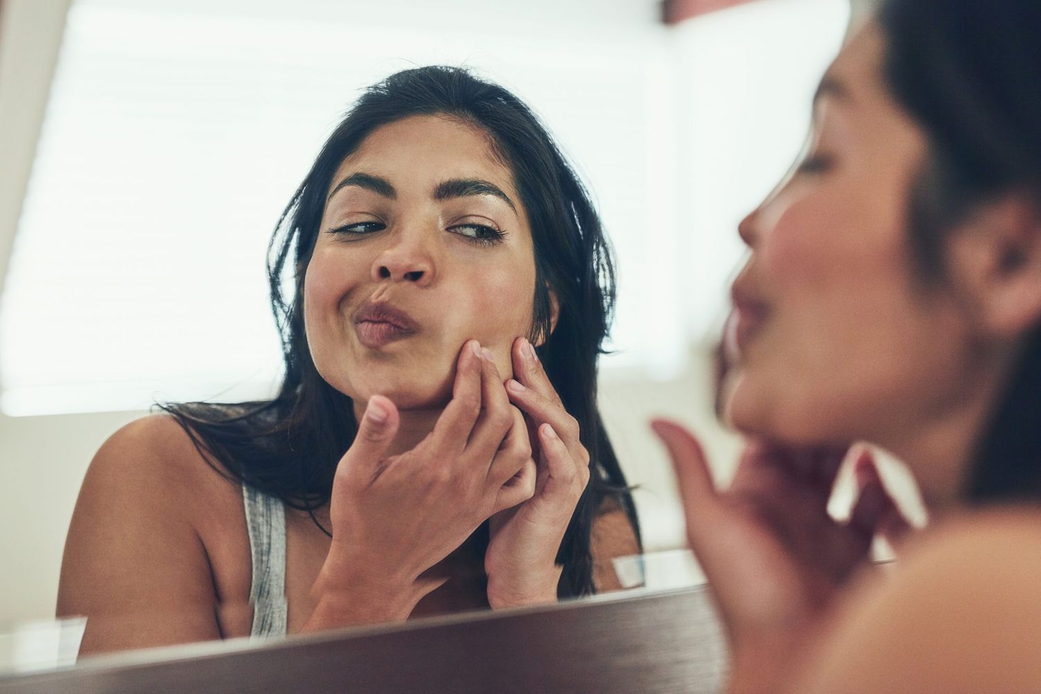 woman popping pimple on face looking in mirror