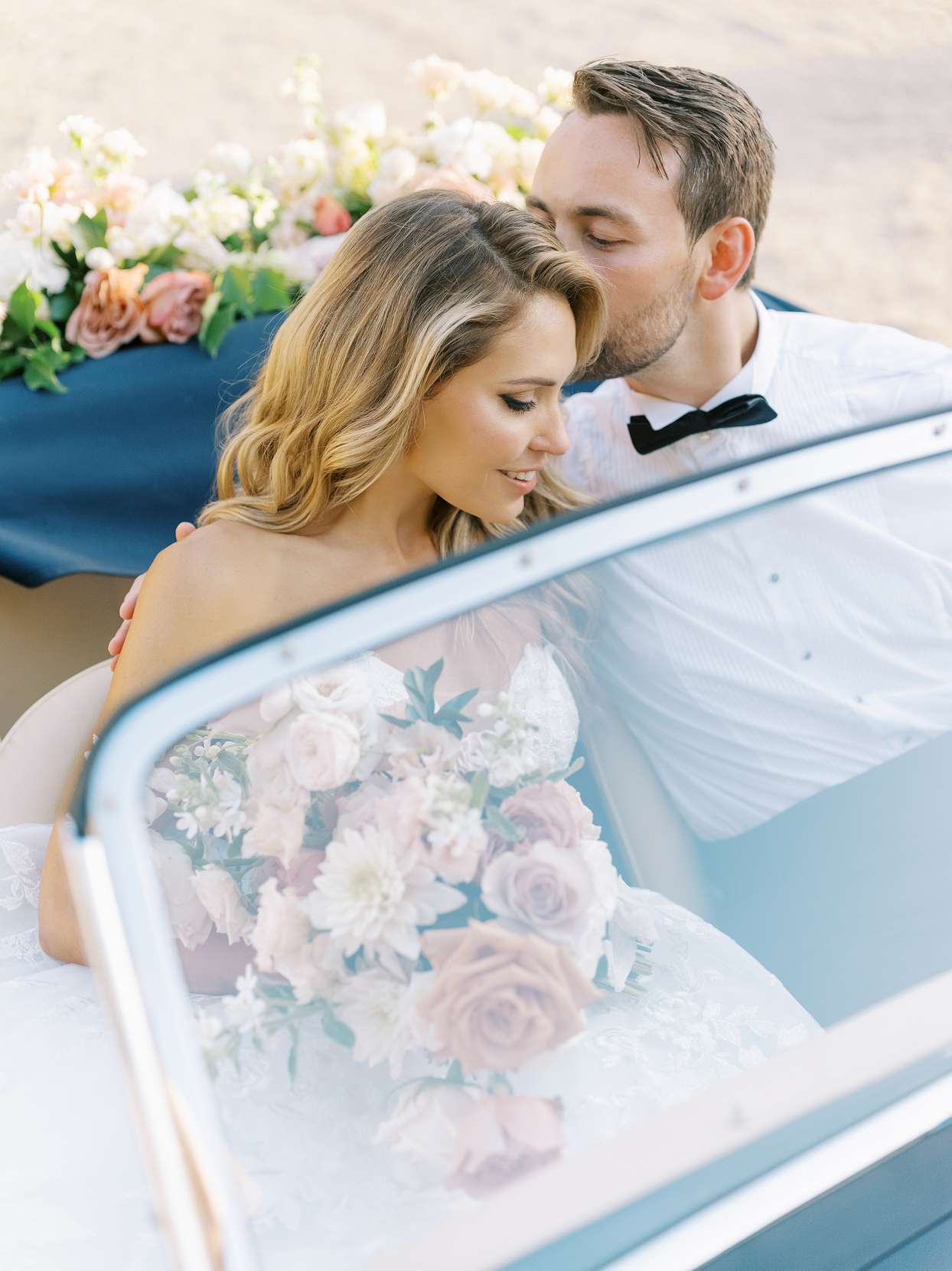 groom kissing bride on head while sitting in black vintage car