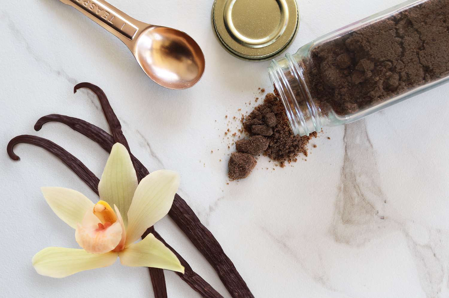 Ground vanilla in a glass bottle, metal teaspoon, and vanilla beans on a marble countertop