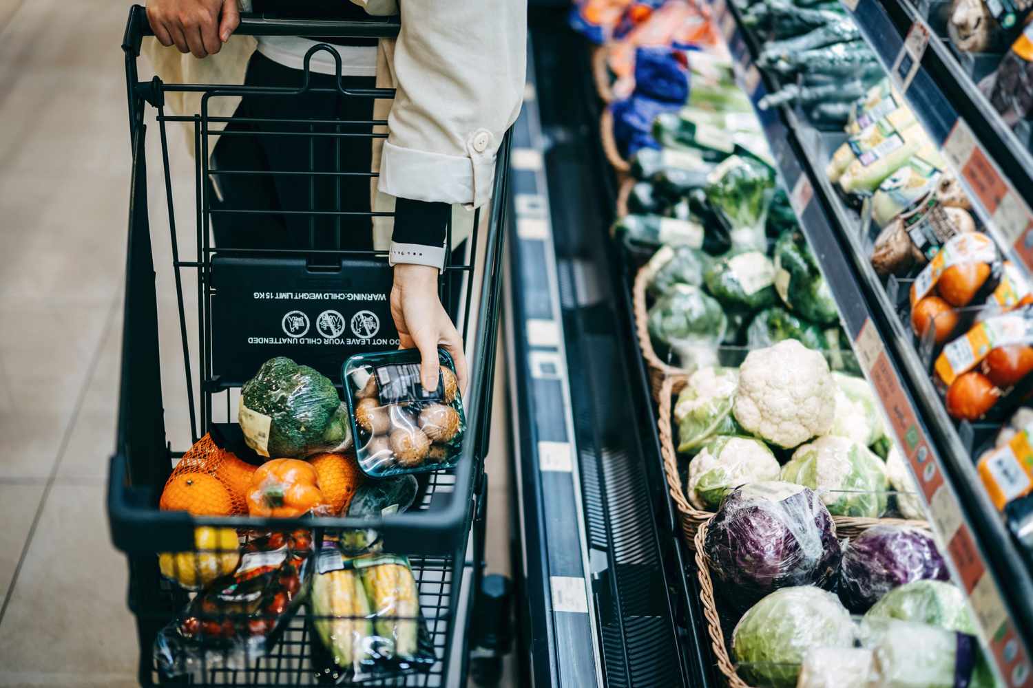 Close up of woman’s hand shopping for fresh groceries in supermarket and putting a variety of organic vegetables in shopping cart