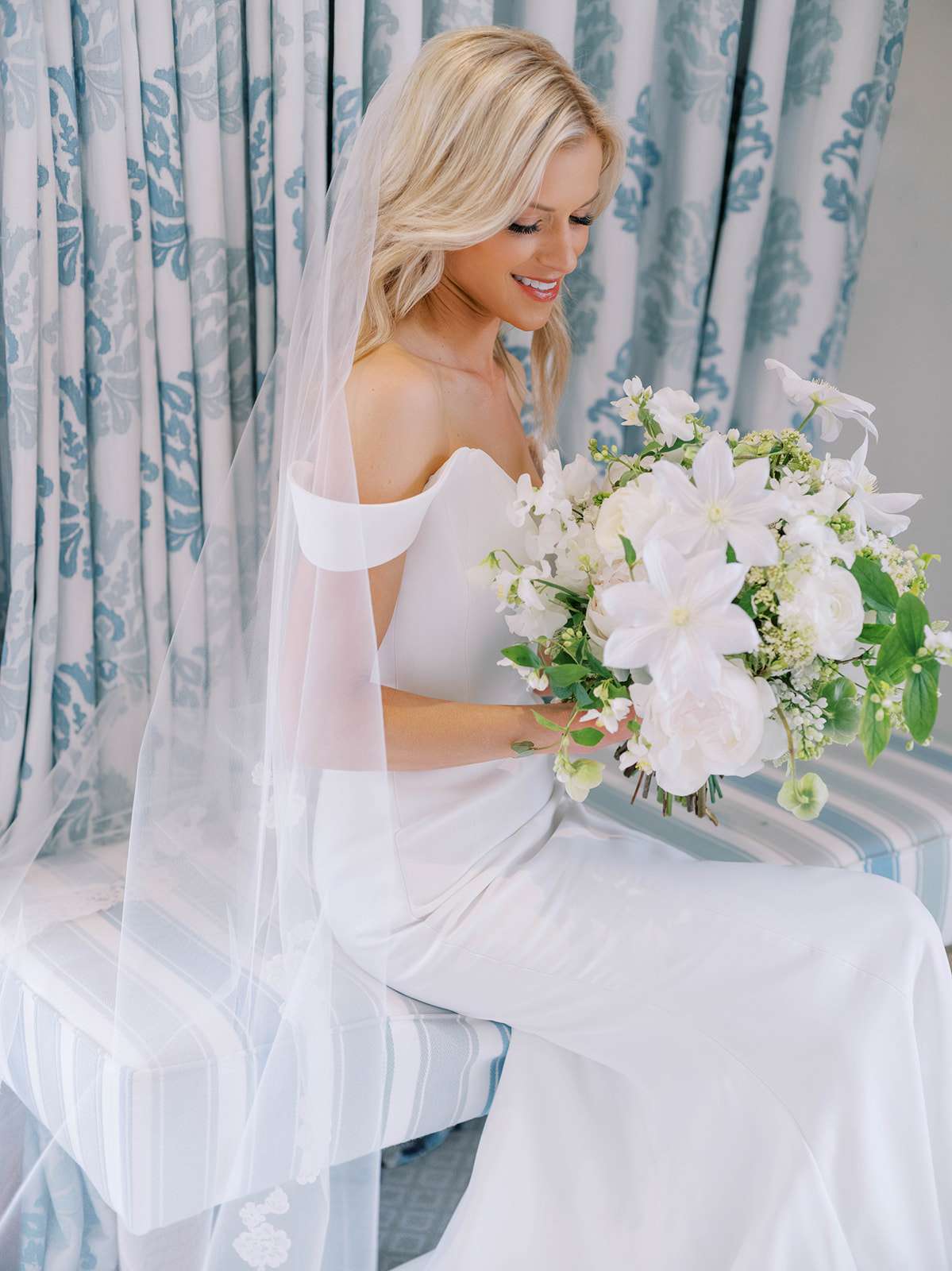 bride sitting in off the shoulder white dress with veil and flowers