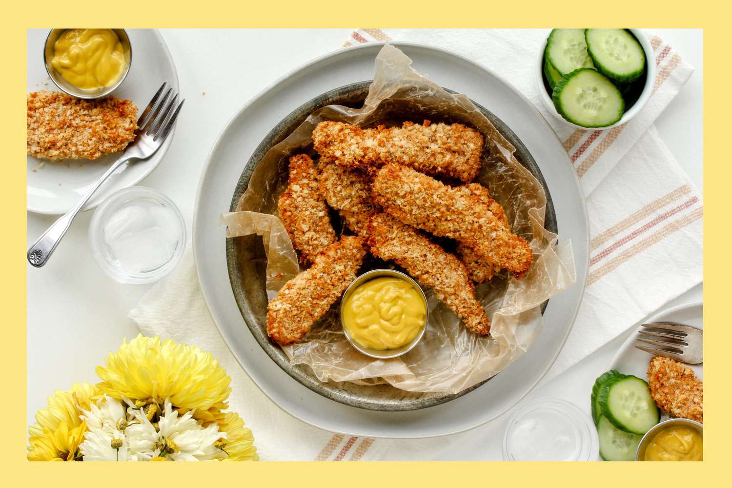 Homemade Chicken Tenders on a serving plate styled with cucumbers, mustard and ketchup on a white background