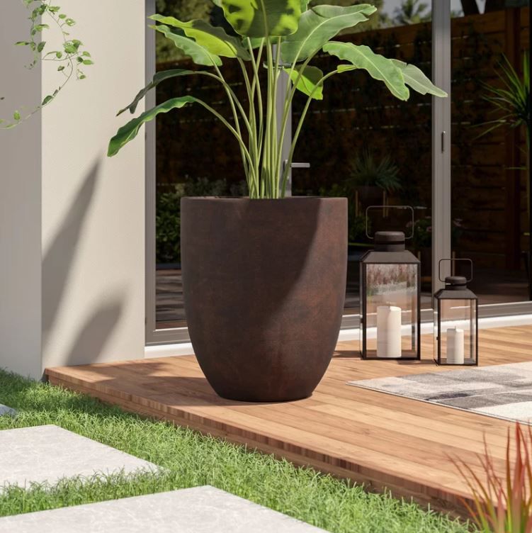 Large planter on wood patio with dark brown, rusted finish and a tropical plant with big leaves inside