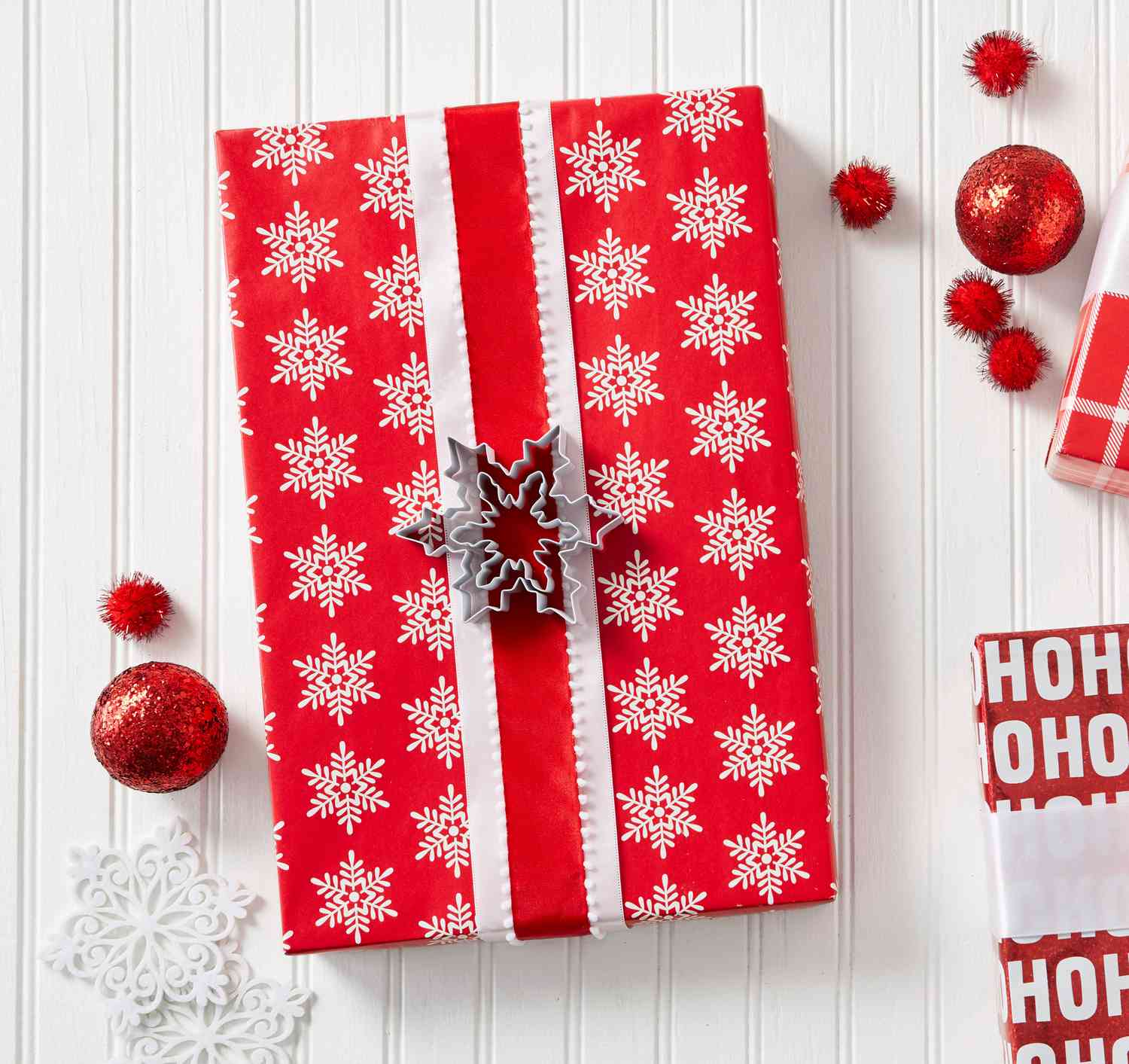 gift box wrapped in red and white snowflake wrapping paper, next to white snowflake ornaments and red sparkly ball ornaments