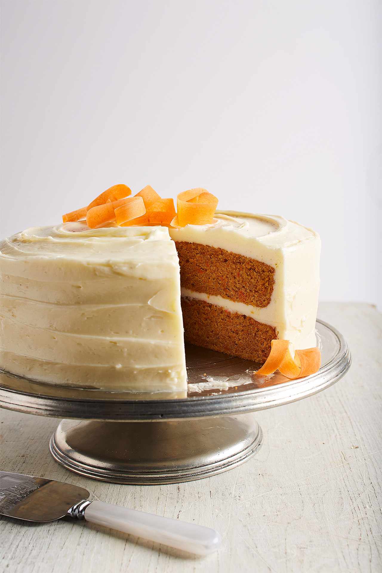 Carrot Cake Recipe on a Silver Cake stand with a slice cut out. Carrot ribbons on top.