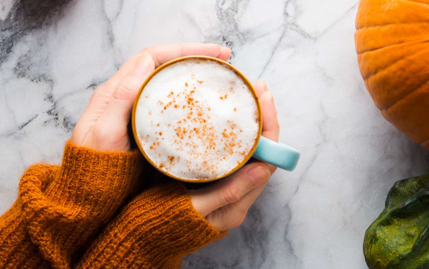 hands holding a pumpkin latte on a marble surface with a pumpkin nearby