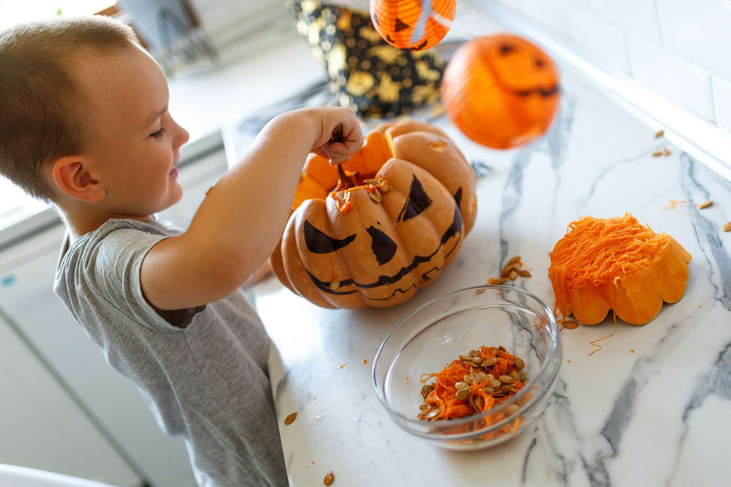 child scooping out pumpkin seeds and insides from a fresh pumpkin with jakc-o-lantern markings on a kitchen counter