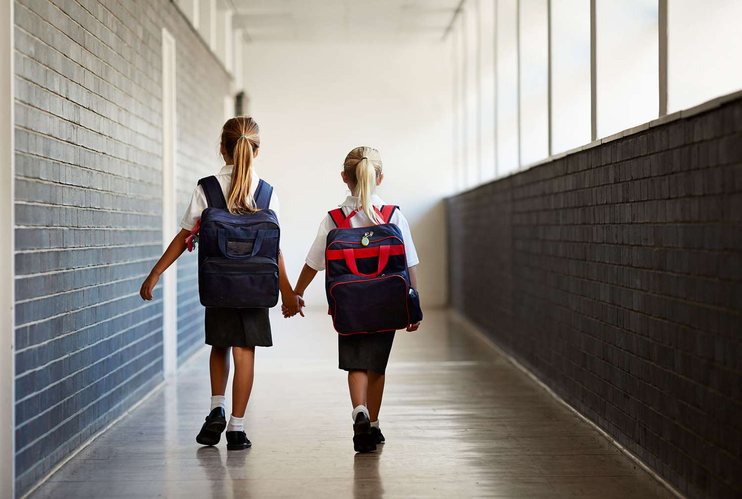 Back of Two School Girls Wearing Backpack Holding Hands Walking Down Hallway