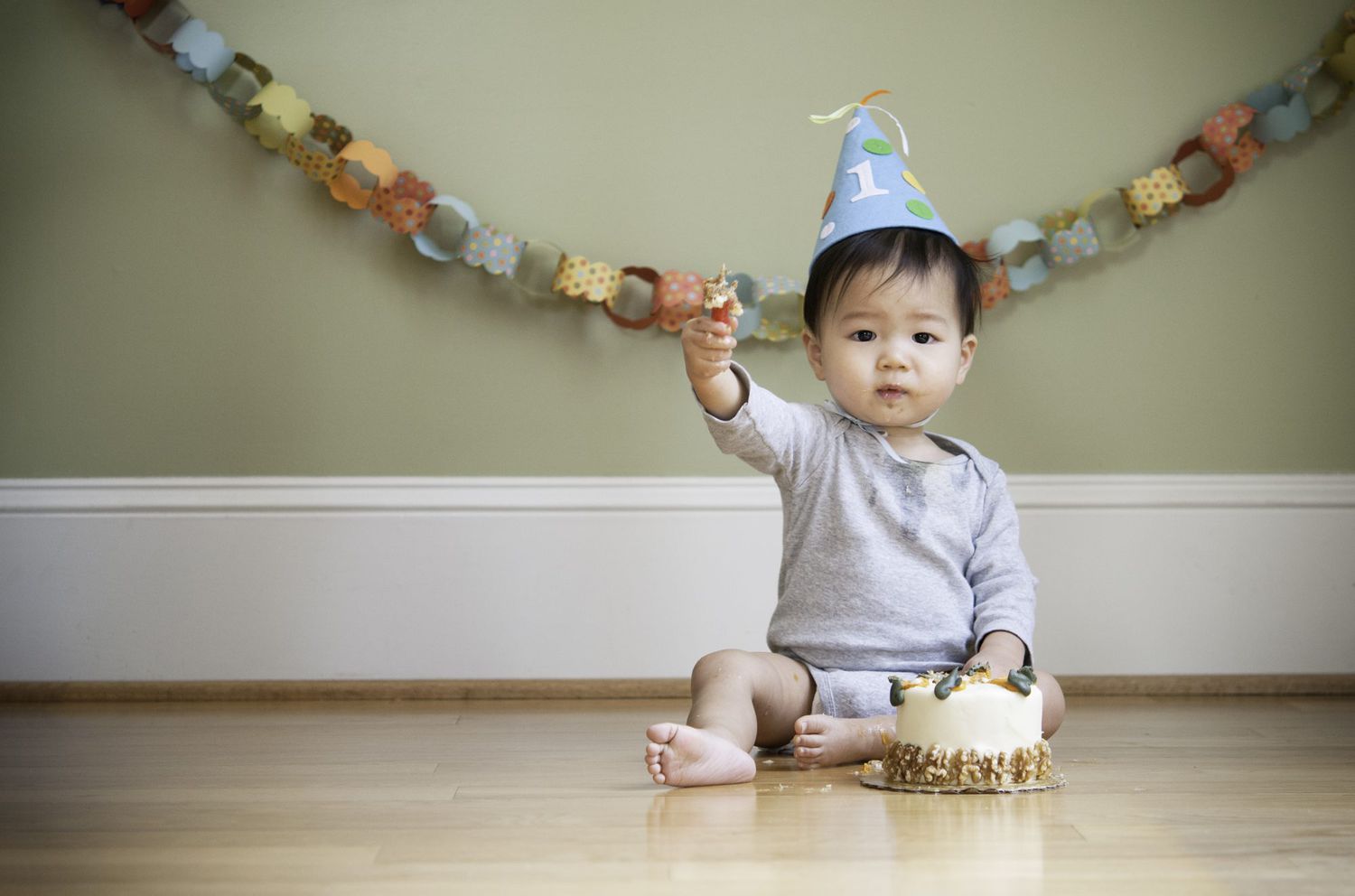 aby celebrating his First Birthday in front of his birthday cake at his birthday cake smash photoshoot. He's holding out the birthday candle to the camera.