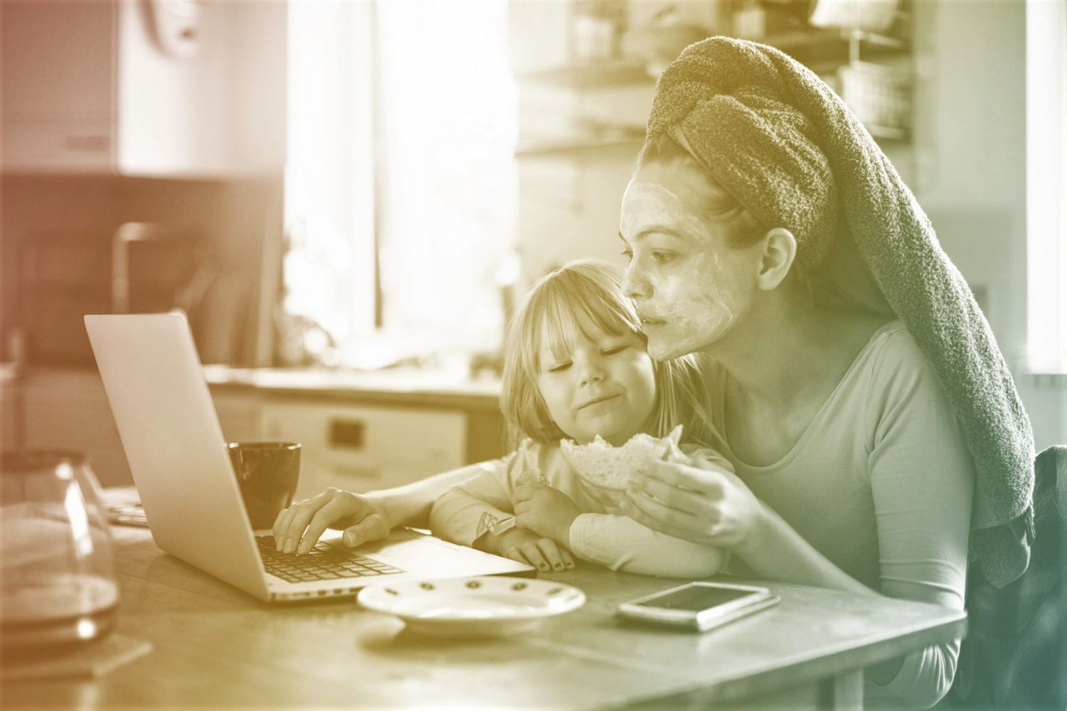 Mother with hair in towel and daughter having breakfast in kitchen while on laptop