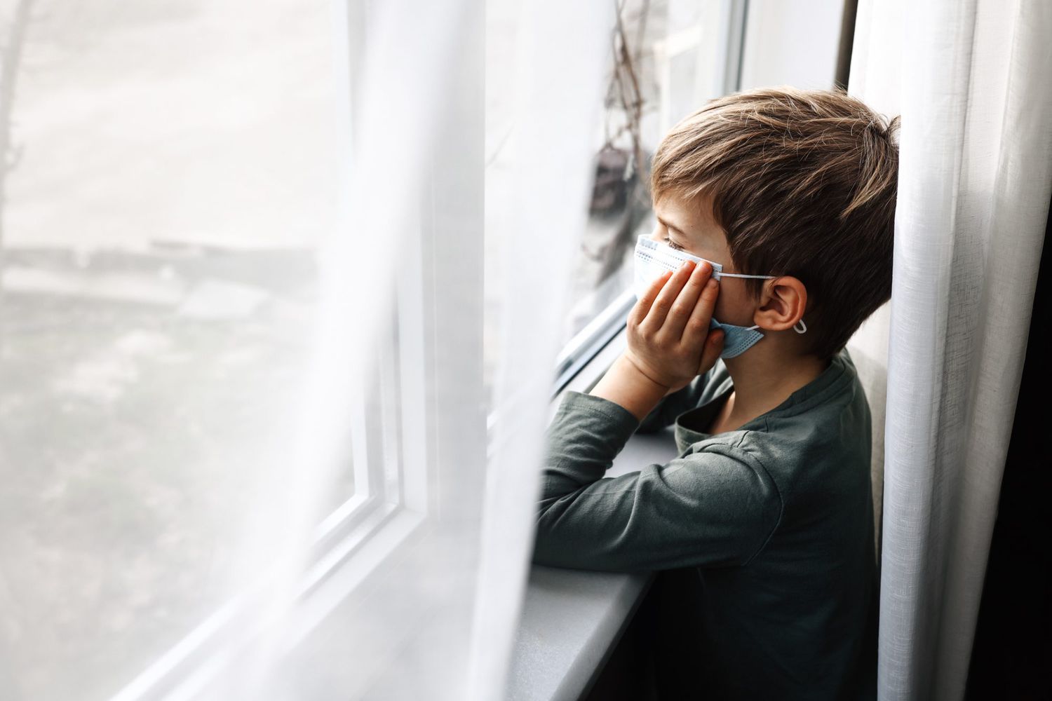 An image of a boy looking out a window with a mask on.